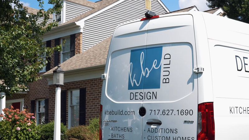 White design-build van parked beside a brick house under construction, with blue logo and phone number.