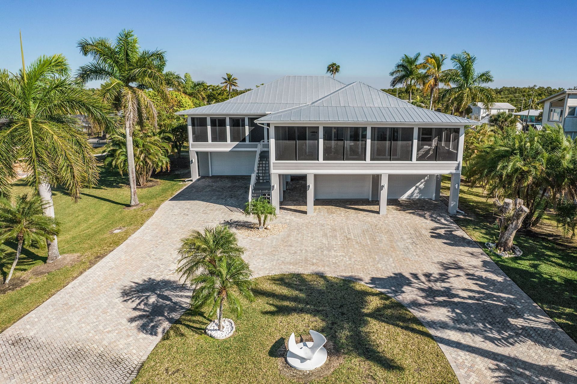 Elevated house with gray roof and screened porch, surrounded by palm trees and gravel driveway under a blue sky.