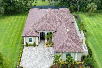 Aerial view of a beige house with a red-tiled roof, set on a green lawn with a stone driveway.