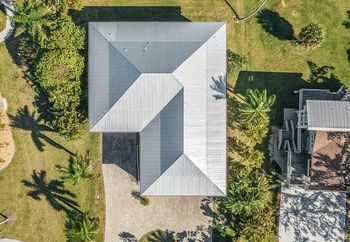 Aerial view of a gray-roofed house with gravel driveway, surrounded by green grass and trees.