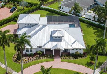 Aerial view of a white modern home with a metal roof, brick driveway, and a swimming pool surrounded by green lawn and palm trees.