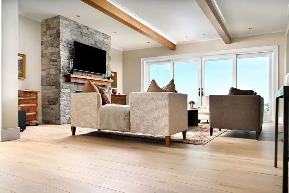 Living room with stone fireplace, TV, beige sofas, large windows, and light wood flooring.
