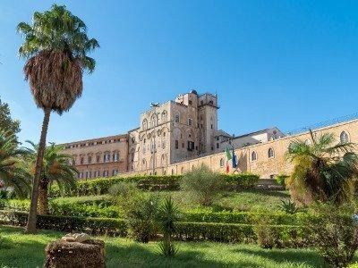 Un grande edificio storico in pietra con torre centrale dietro un giardino paesaggistico con siepi e palme ben curate.