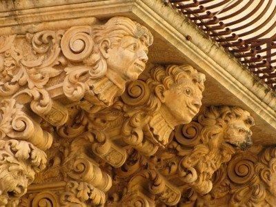 Elaborate sculture in arenaria raffiguranti volti e volute floreali ornate che sostengono un balcone su un edificio.
