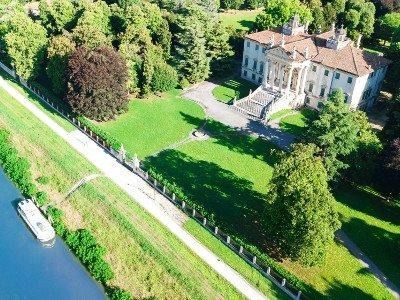 Una vista aerea di una villa con tetto in tegole, circondata da prati verdi e alberi, accanto a un fiume con una barca.