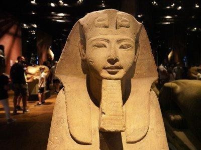 Una statua di sfinge con il tradizionale copricapo nemes e la barba finta esposta in un museo scarsamente illuminato.
