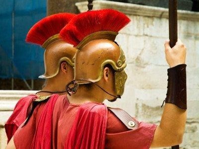 Due persone vestite da soldati romani, con tuniche rosse ed elmi con creste rosse di profilo e impugnano delle lance.