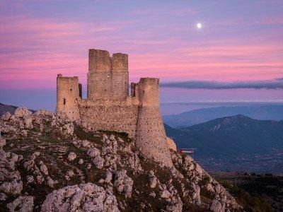 Rovine di un castello  sulla cima di una montagna rocciosa al tramonto, con una luna fioca in un cielo rosa e viola.