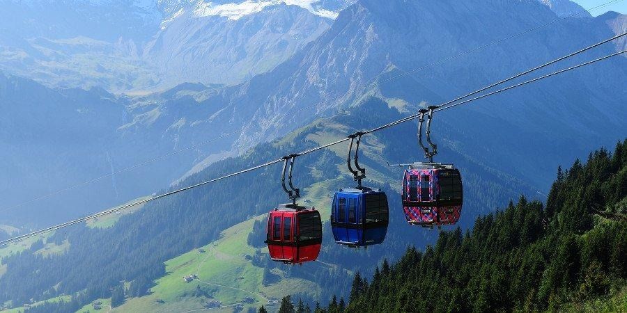 Le cabine rosse, blu e viola scorrono lungo un cavo sopra un pendio montuoso boscoso con cime innevate sullo sfondo.