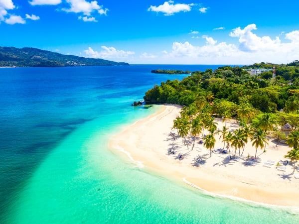 Vista di una spiaggia di sabbia bianca con acqua turchese, palme e una lussureggiante foresta verde sotto un cielo azzurro.