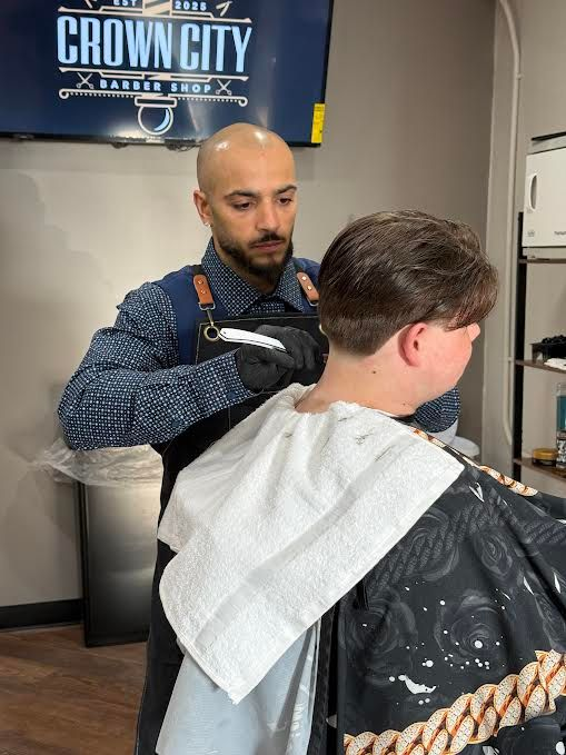 Barber trimming a customer’s hair in a shop, with a CROWN CITY sign on the wall.