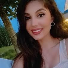 Smiling woman with long dark hair and hoop earrings outdoors at sunset, with greenery behind her
