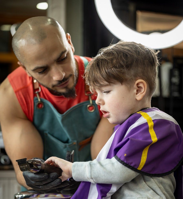 Barber showing a child how to hold scissors in a salon, both focused on a haircut.