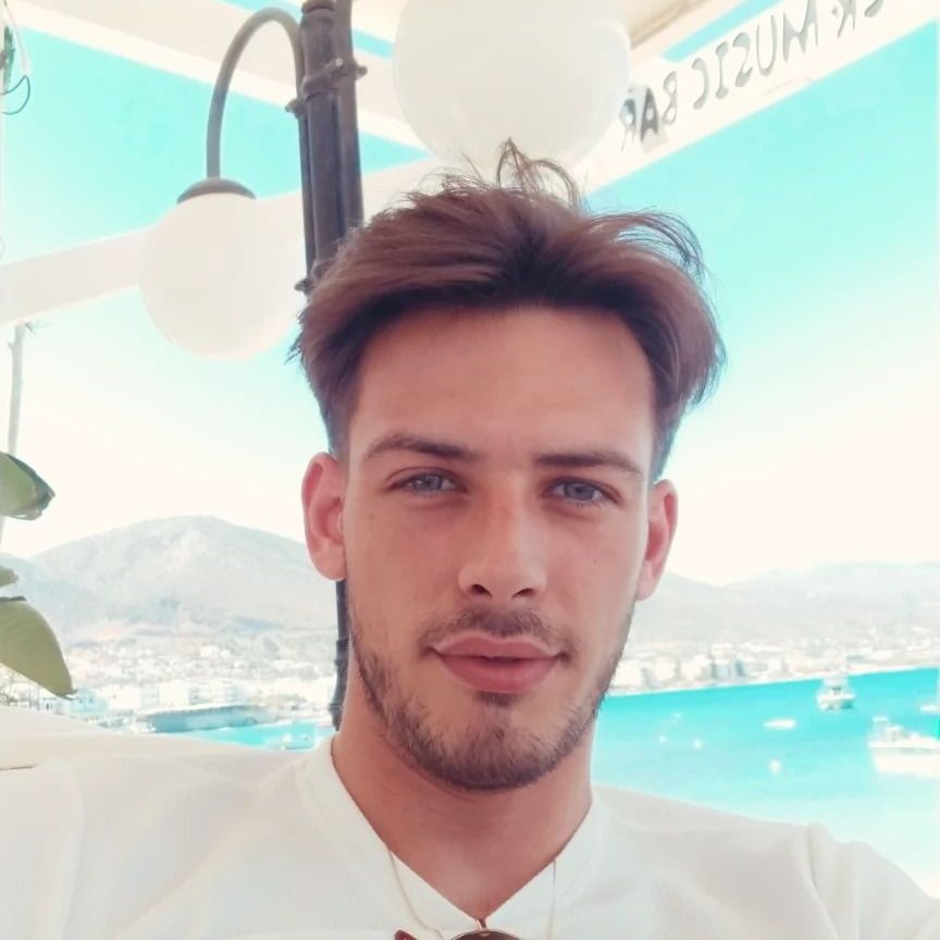 Een man met bruin haar en stoppels glimlacht, zittend in de buitenlucht met uitzicht op een blauwe zee.