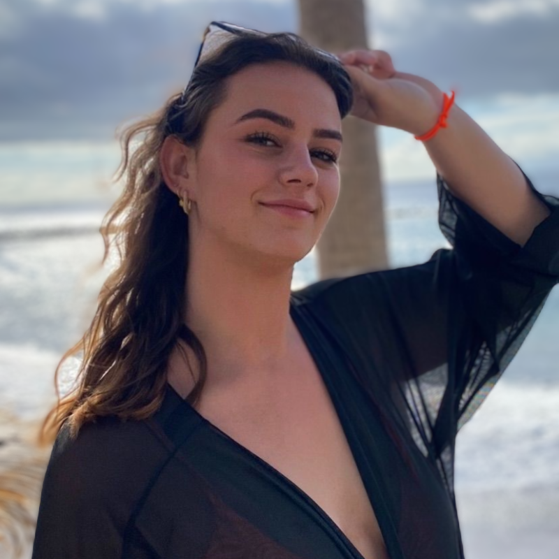 Vrouw in een zwarte omslagdoek lacht naar de camera, met een zonnebril op haar hoofd. Strand en oceaan op de achtergrond.