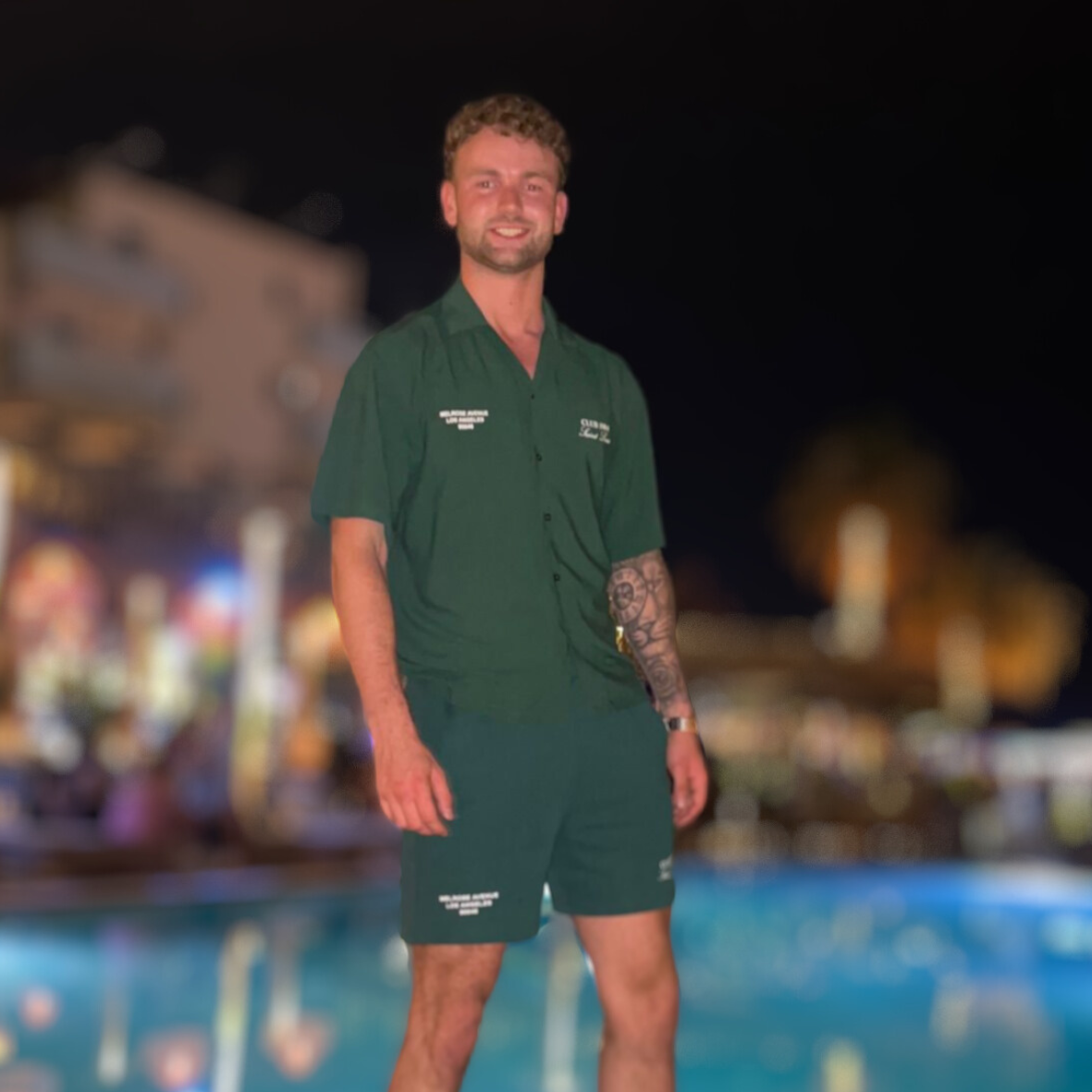 Een man in een groene outfit staat 's nachts bij een zwembad en glimlacht.