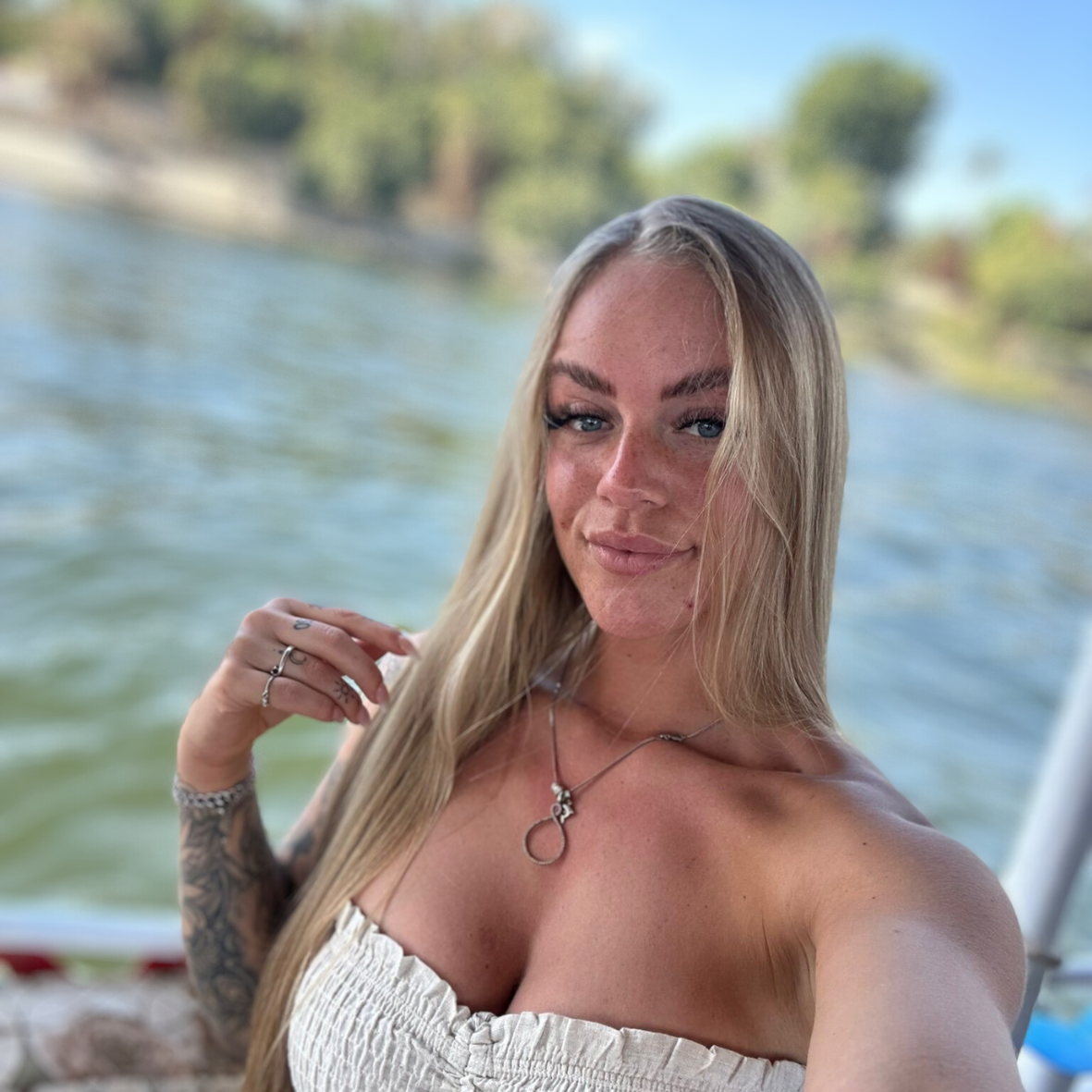 Vrouw met lang blond haar, een strapless topje en sieraden, lacht naar de camera vanaf een boot met water op de achtergrond.