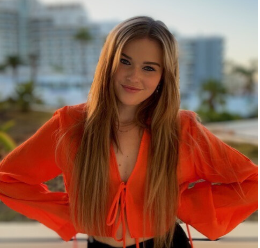 Vrouw in oranje top met lang blond haar lacht. Buitenachtergrond.