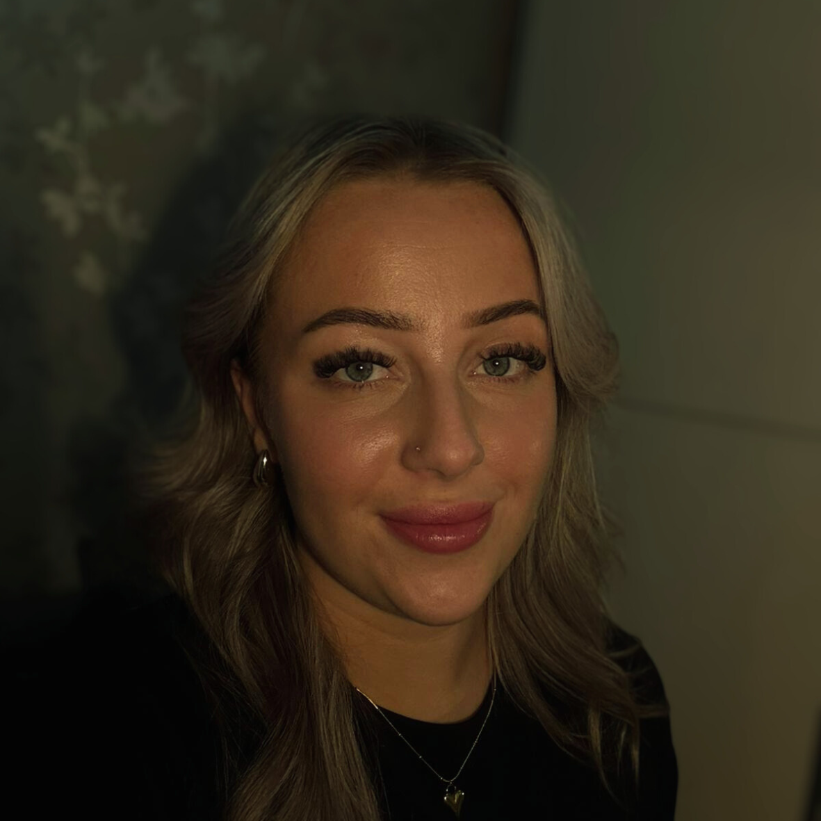 Blonde vrouw met een lichte huid, gekleed in een zwarte blouse en met een ketting om, kijkt in de camera.