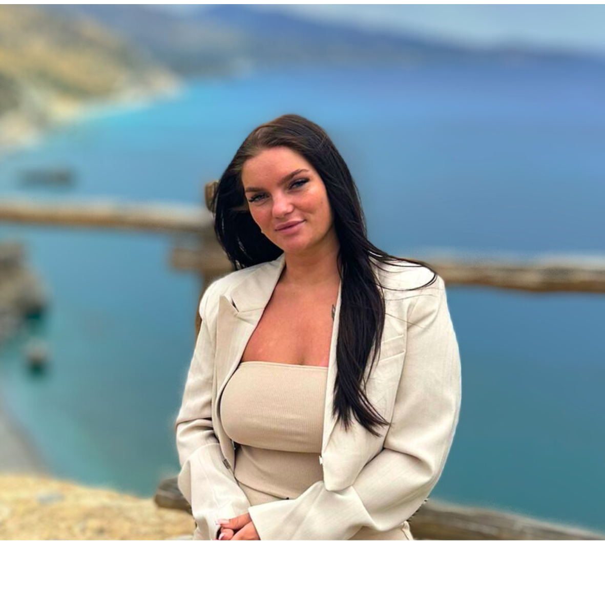 Vrouw in beige blazer en jurk, lachend, met uitzicht op de oceaan en de bergen.