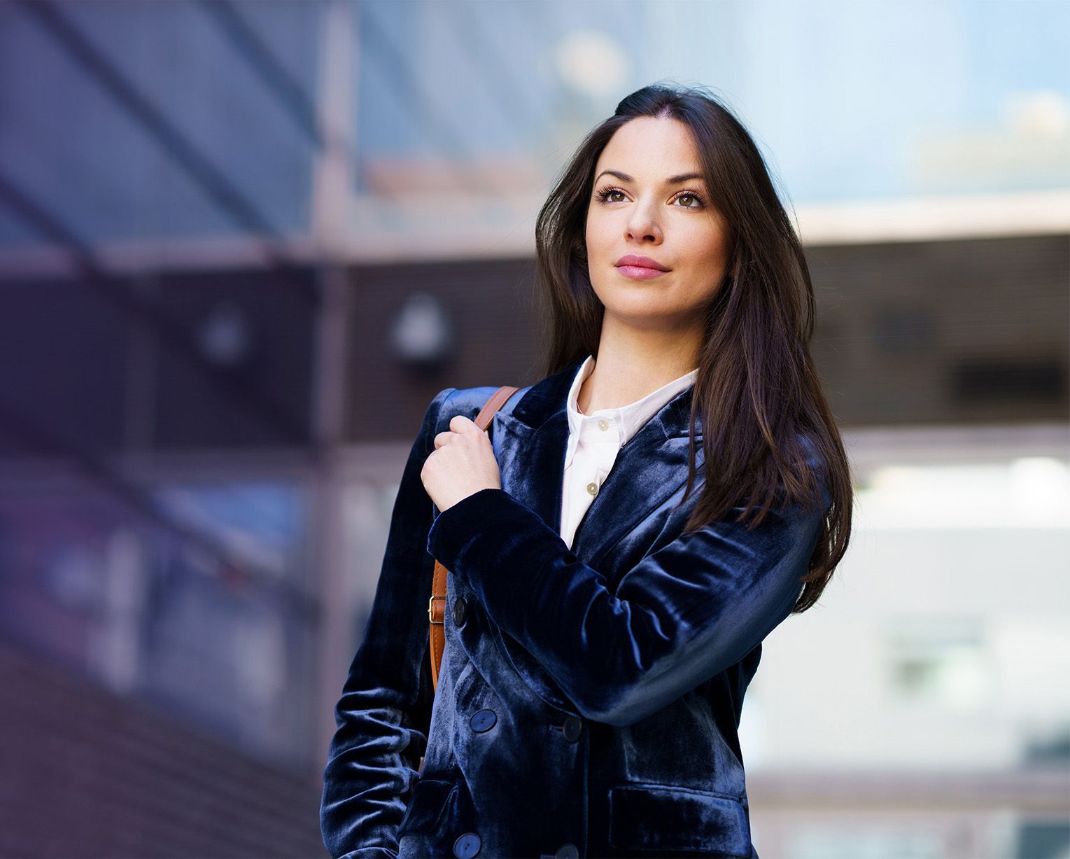 Woman in a dark blue velvet jacket standing outdoors, looking ahead with a shoulder bag