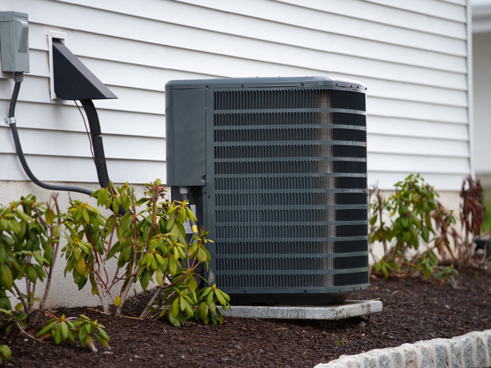 Air conditioning unit outside a building, gray and rectangular, near green shrubs.
