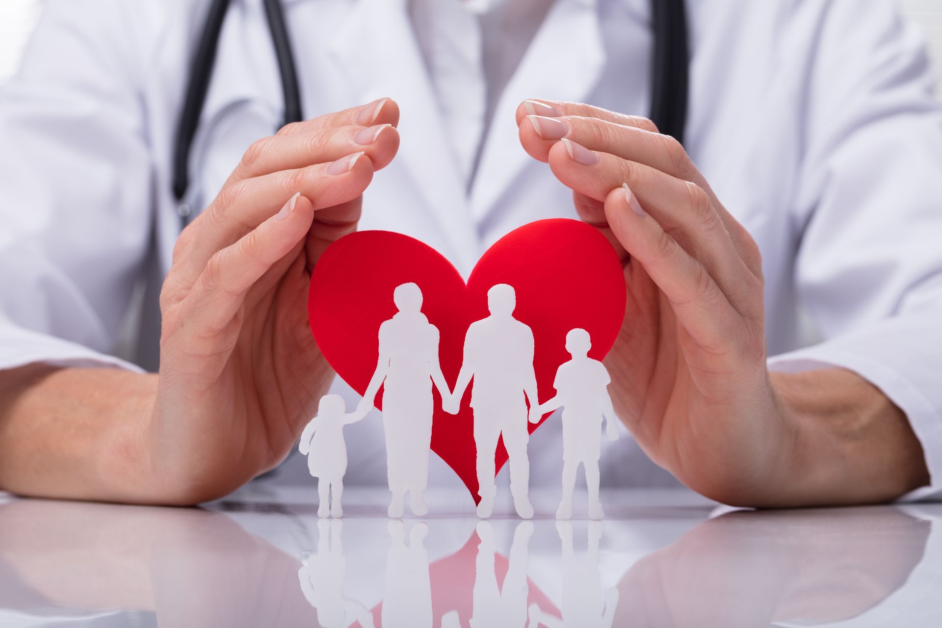 Hands protecting red heart with white family silhouettes on a desk, healthcare concept
