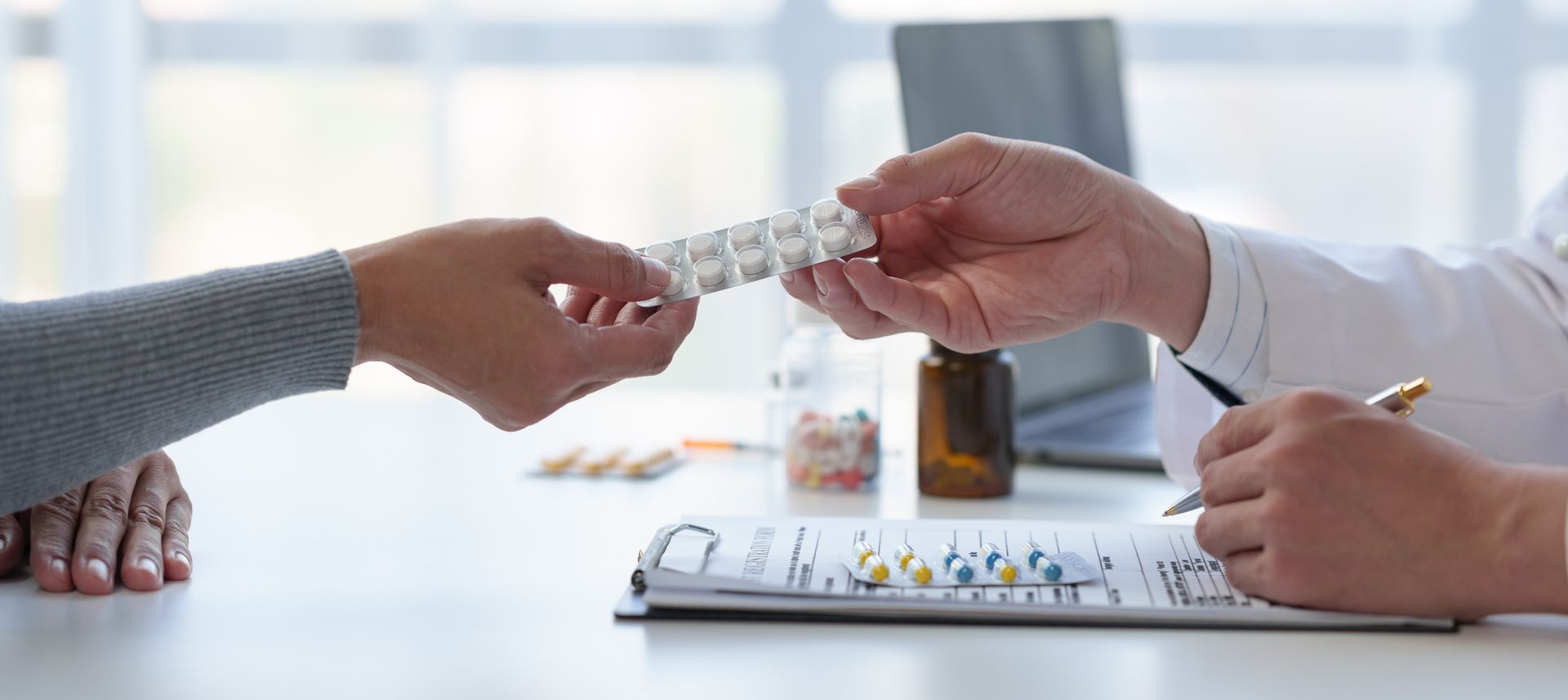 Two hands exchanging a medication blister pack at a desk with pills and a clipboard in the background