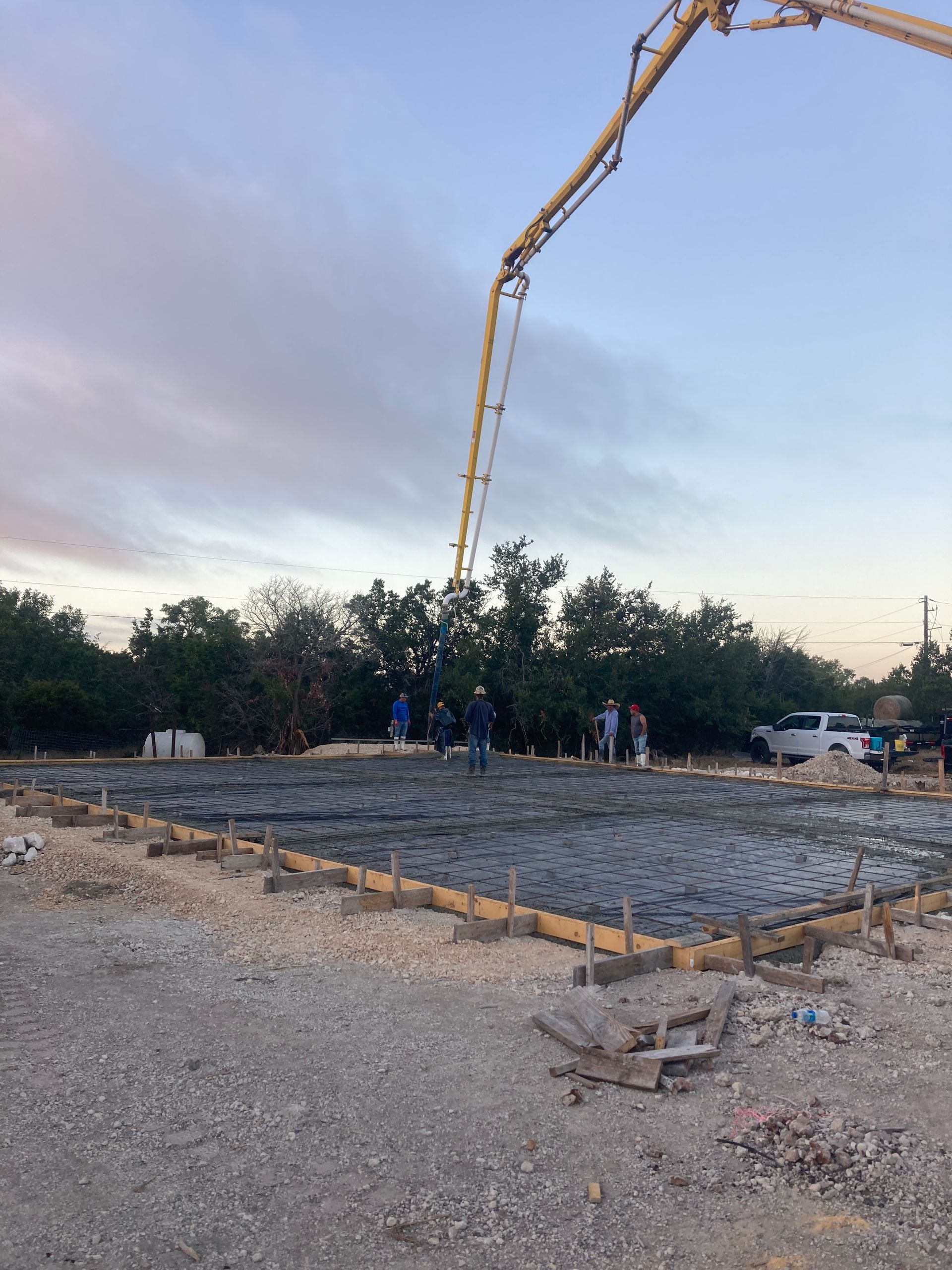 Concrete foundation being poured at dusk by a crane, with workers standing nearby.