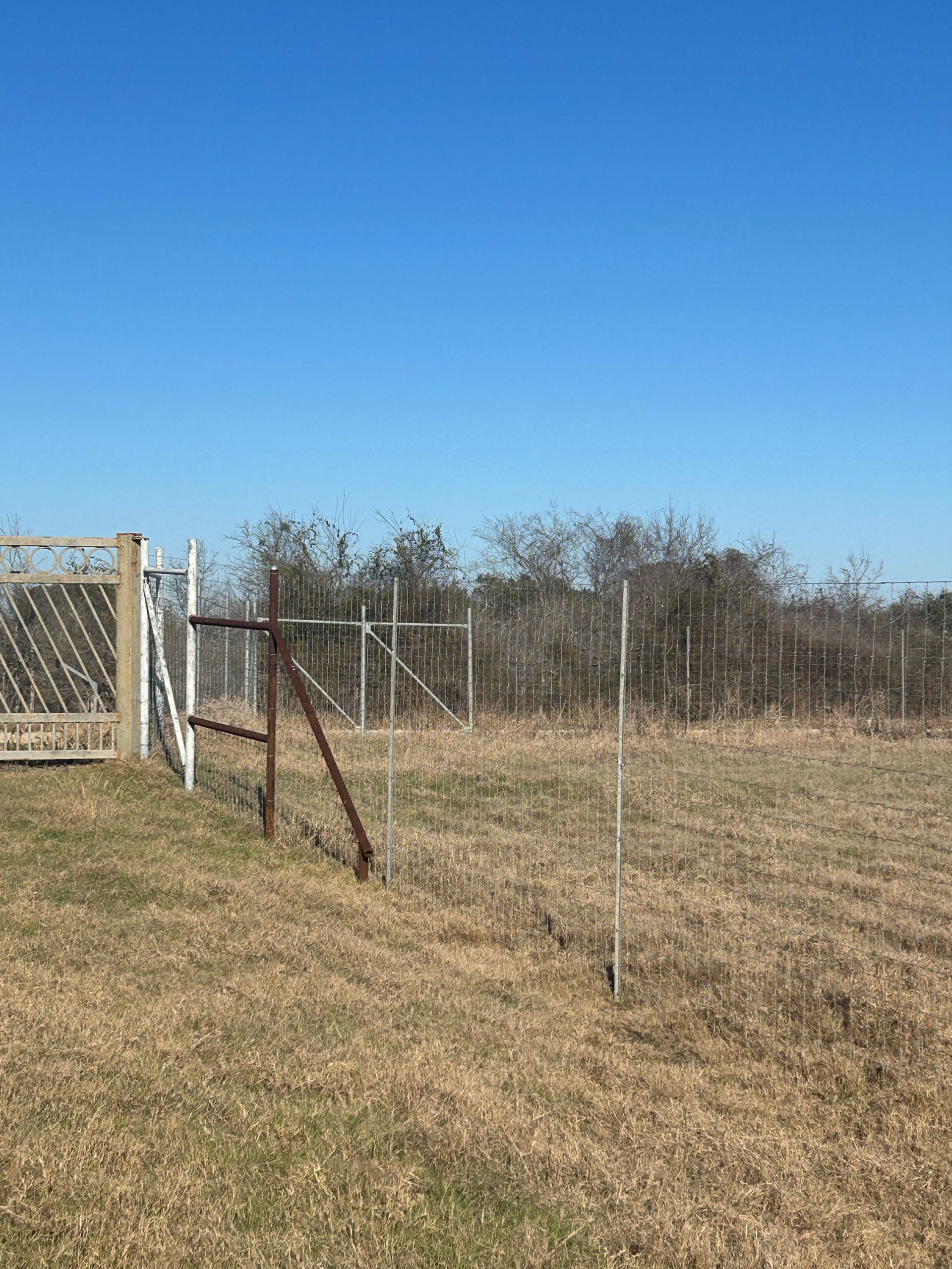 Rusty wire fence beside a gravel road in a dry, open field under a clear blue sky