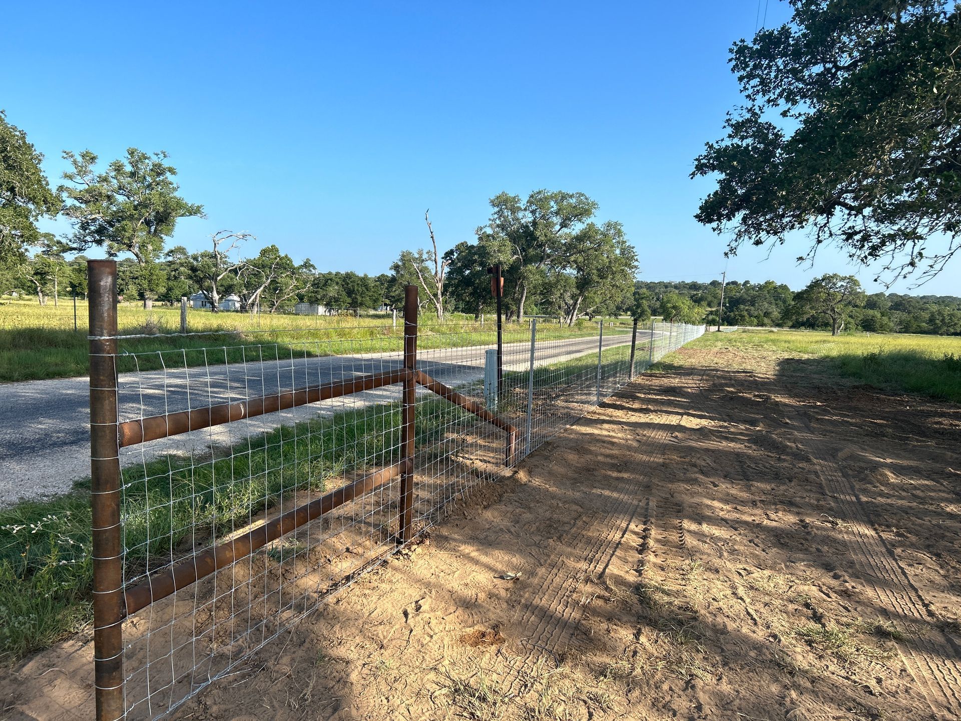 Rusty wire fence beside a dirt road in a sunny rural field with scattered trees