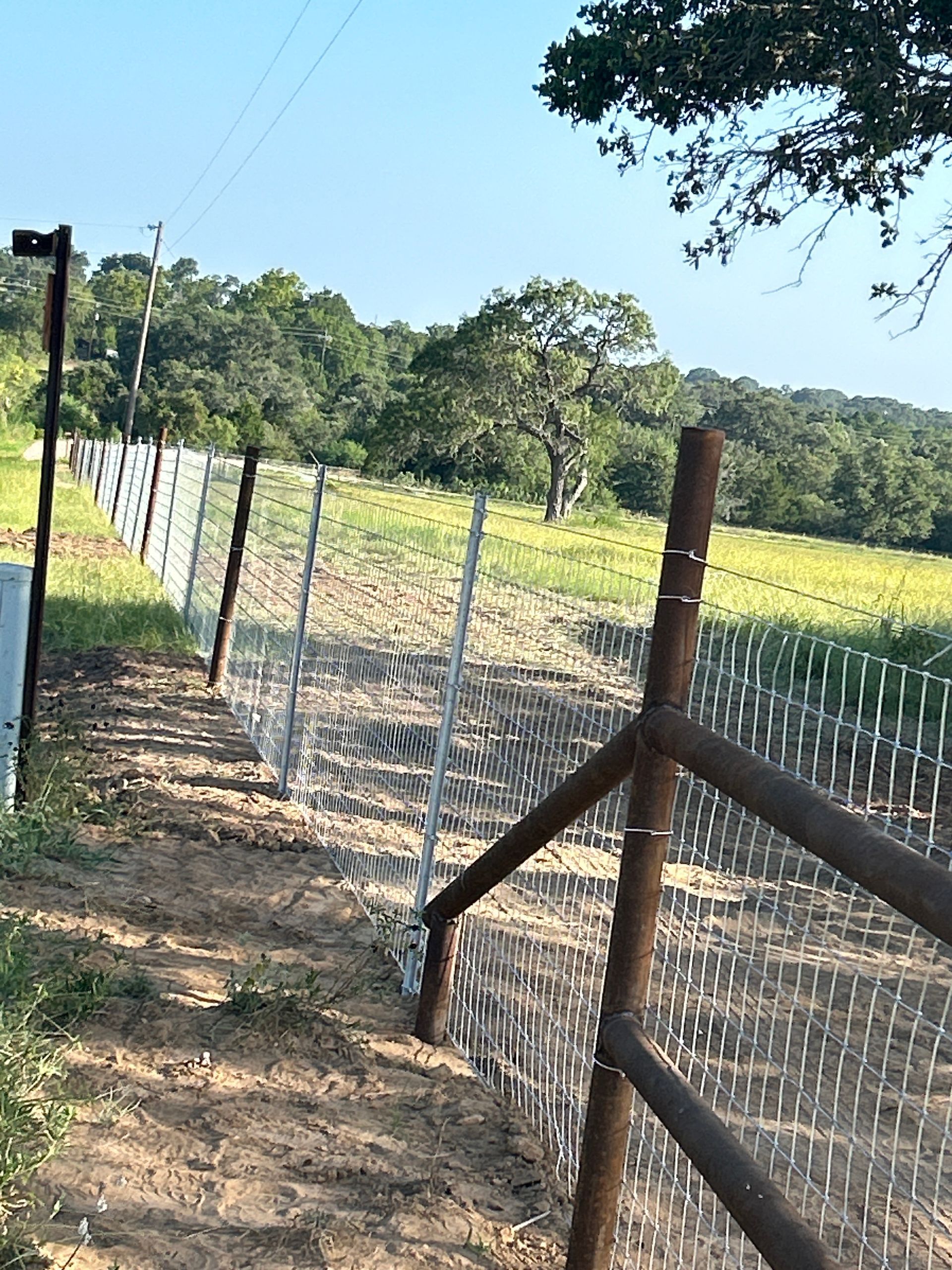 Wire fence along a dirt path beside a green field under a clear blue sky