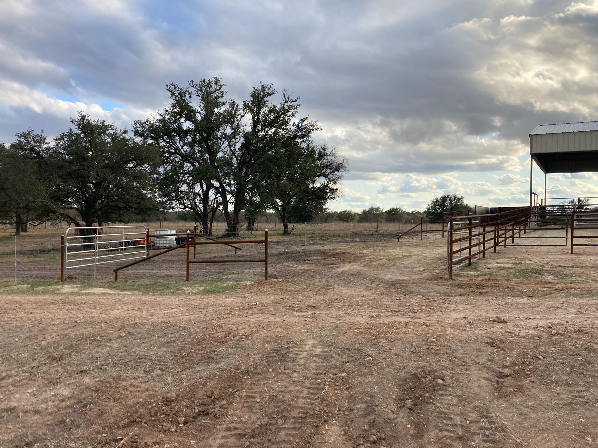 Rural dirt corral with metal gates and trees under a cloudy sky