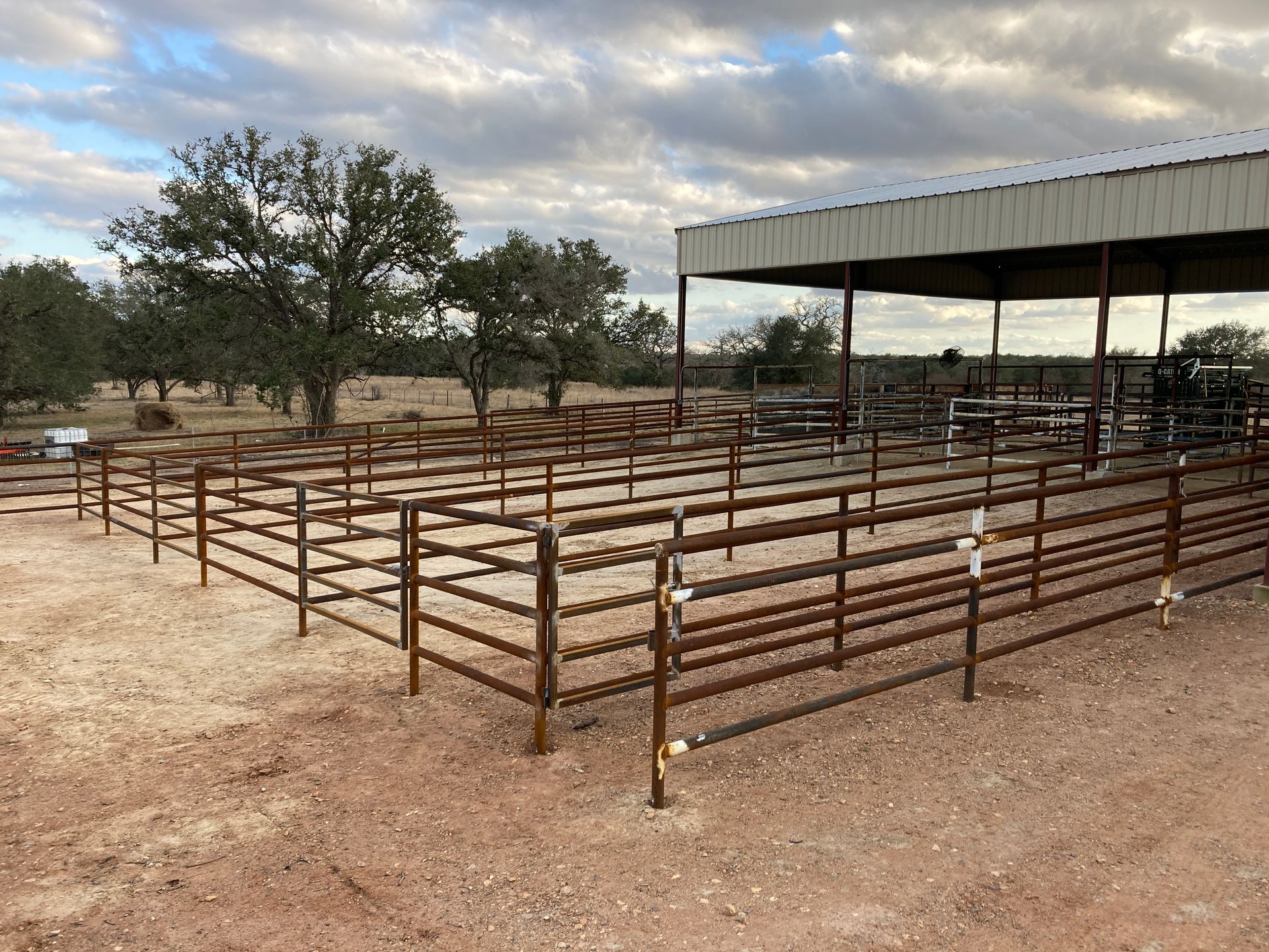 Empty livestock pens with metal fencing beside a covered shed under a cloudy sky.