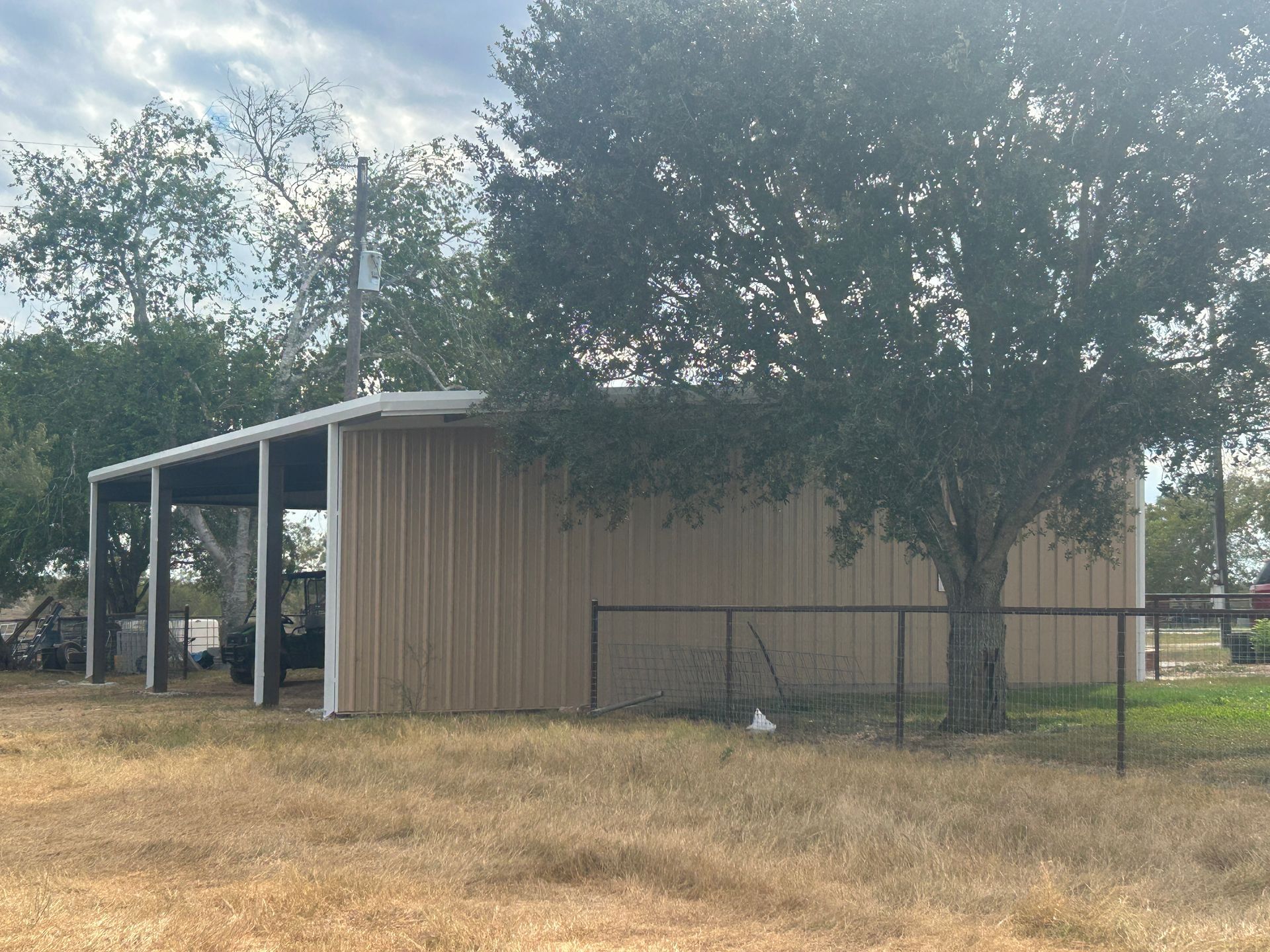Tan metal barn with open shed on a dry grassy lot, framed by trees and a fence