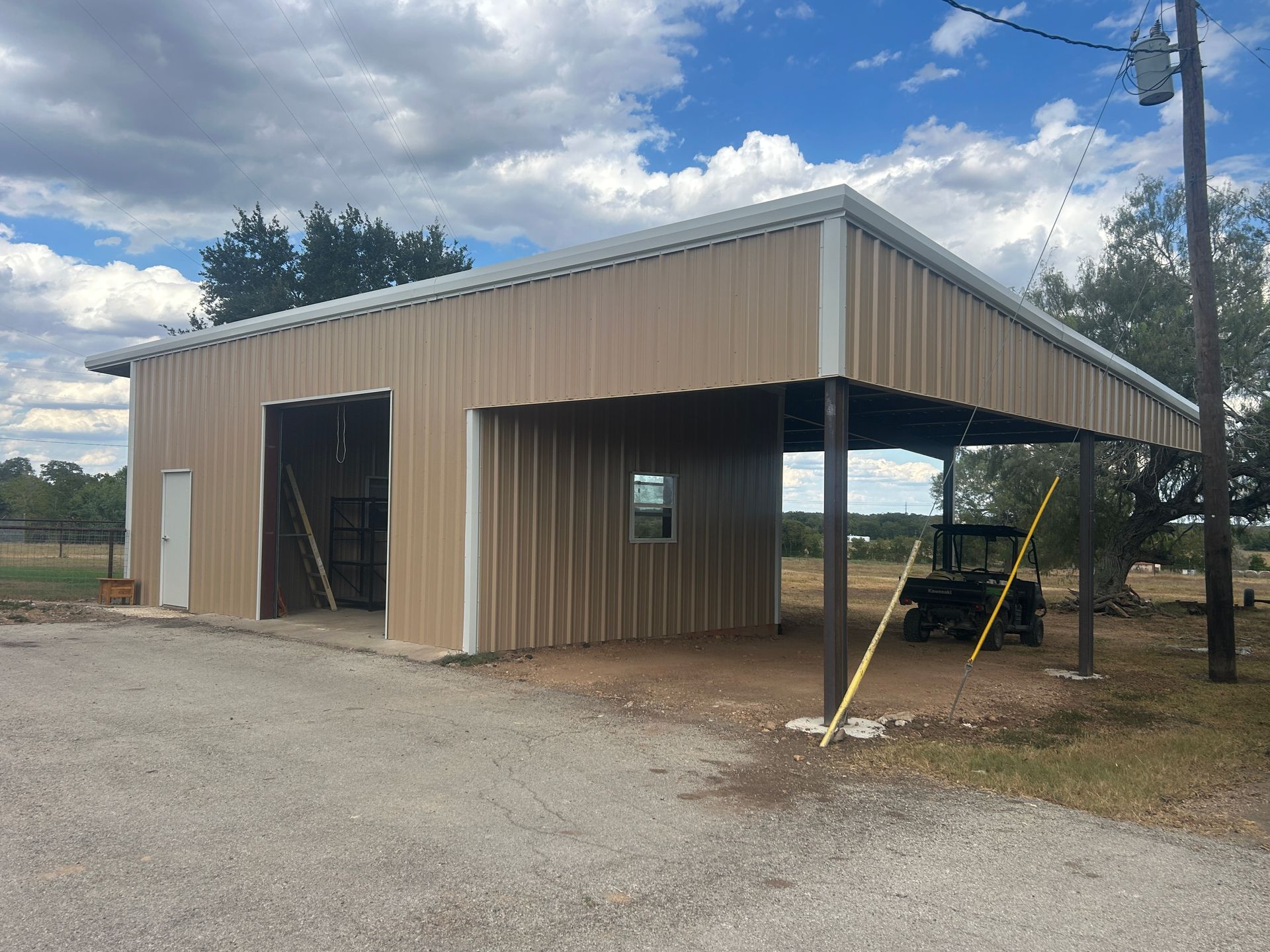 Beige metal workshop building with a covered open bay, set on a gravel lot under a cloudy sky
