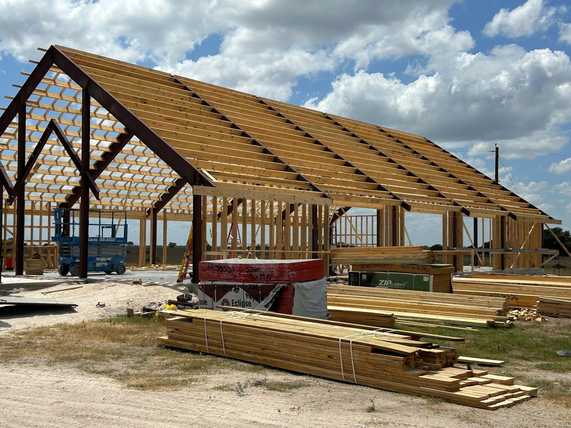Wood-framed house under construction with trusses, lumber stacks, and cloudy sky