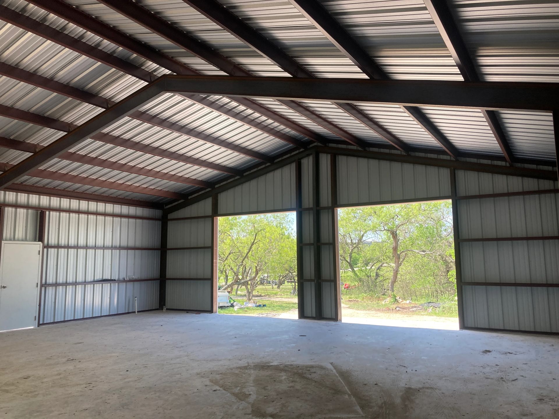 Empty metal garage with open double doors, concrete floor, and trees visible outside.