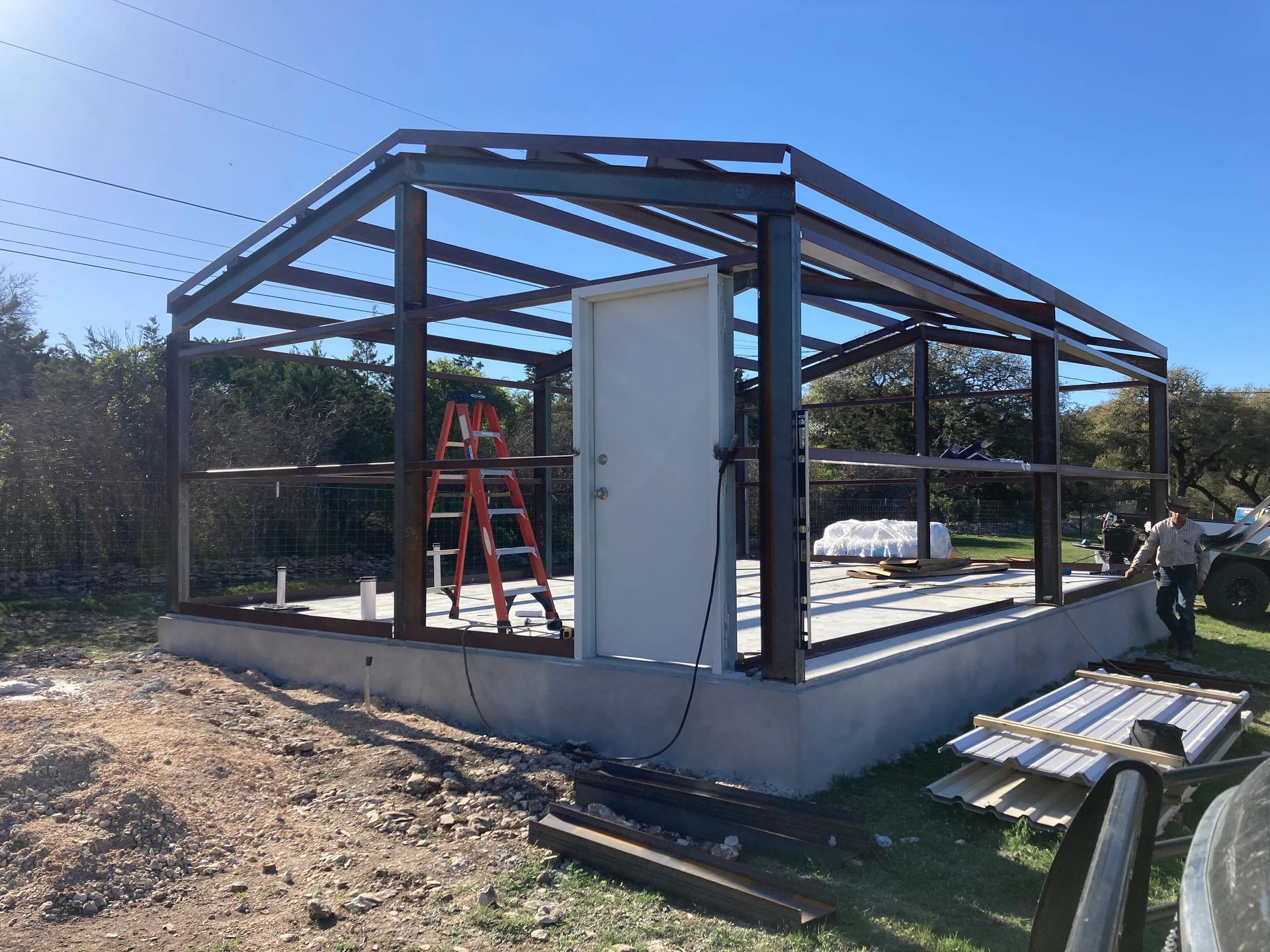 Partially built metal frame shed on a concrete slab, with a red ladder and tools in a sunny yard