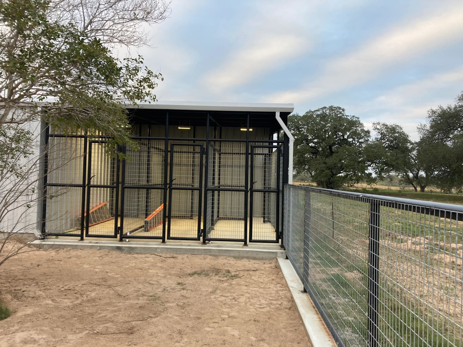 Small black animal enclosure beside a dirt path, with metal fencing and trees in the background.
