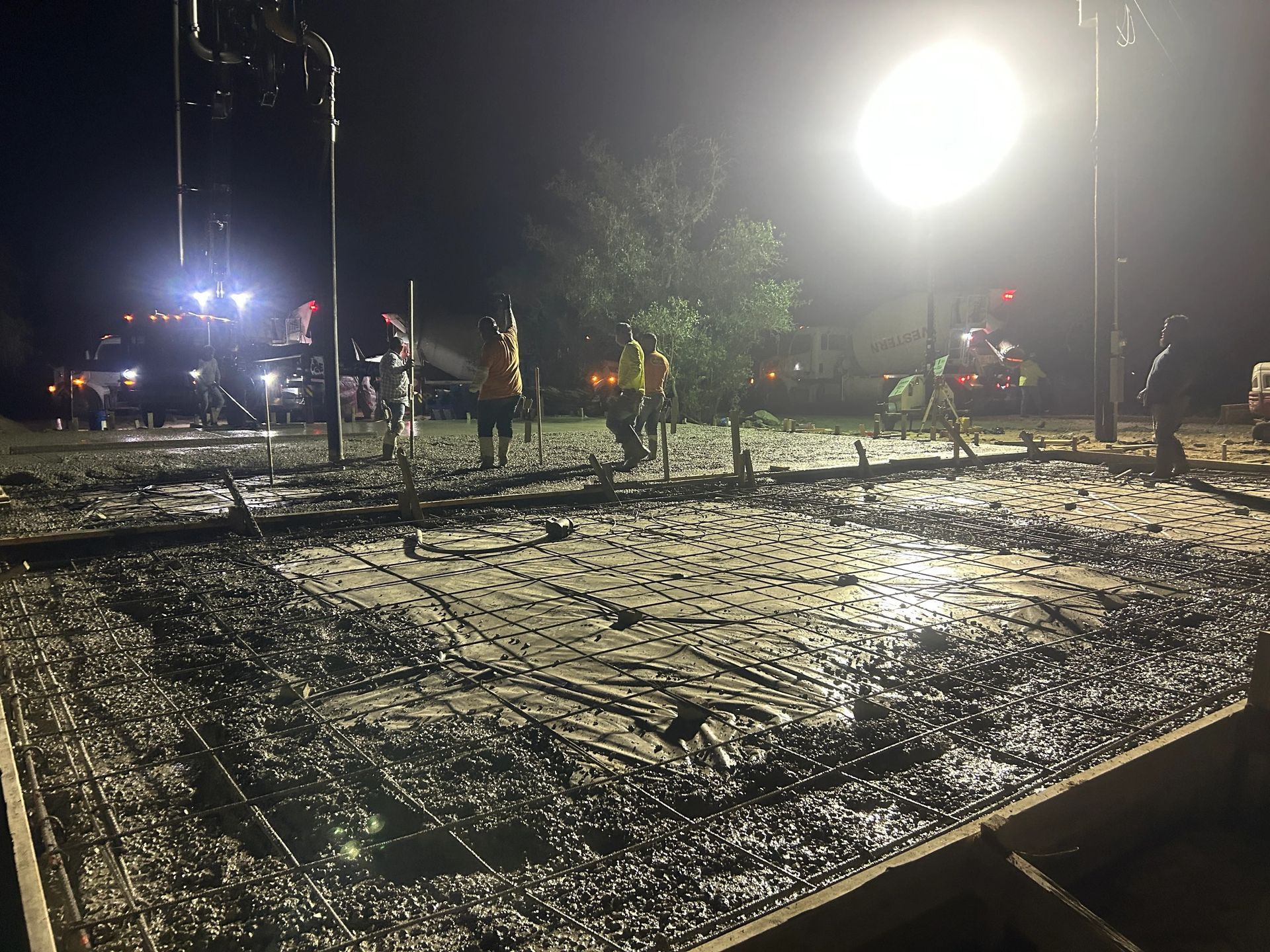 Floodlit muddy construction site at night with workers and heavy equipment in the background