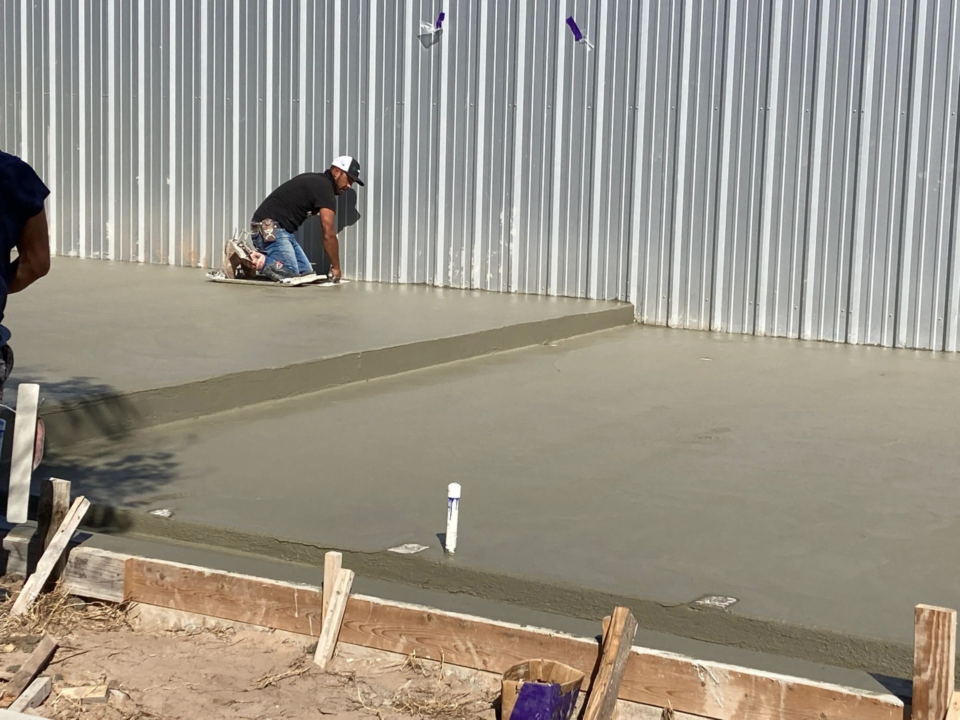 Worker smoothing wet concrete beside a corrugated metal wall at a construction site