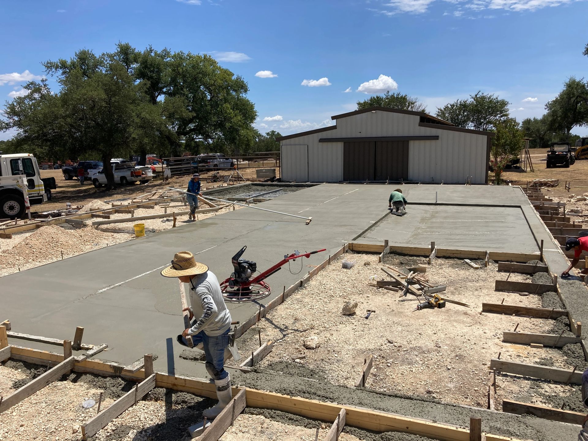 Construction workers pouring a concrete slab beside a metal building under a sunny sky.