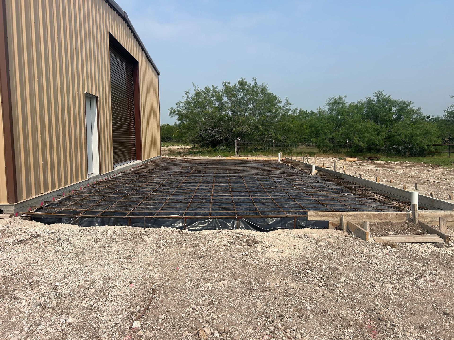 Concrete foundation with rebar beside a metal building, on a gravel construction site.