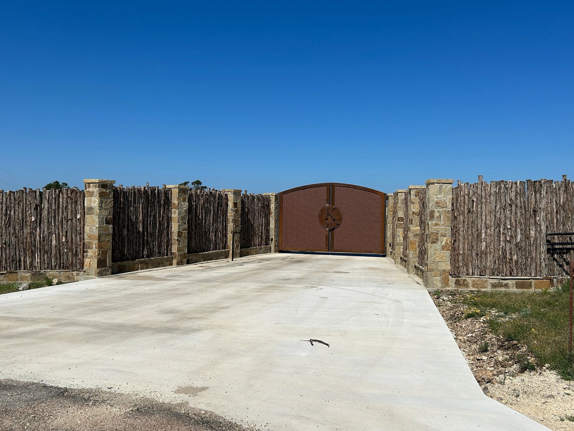 Concrete driveway leading to a closed brown gate between stone walls under a clear blue sky