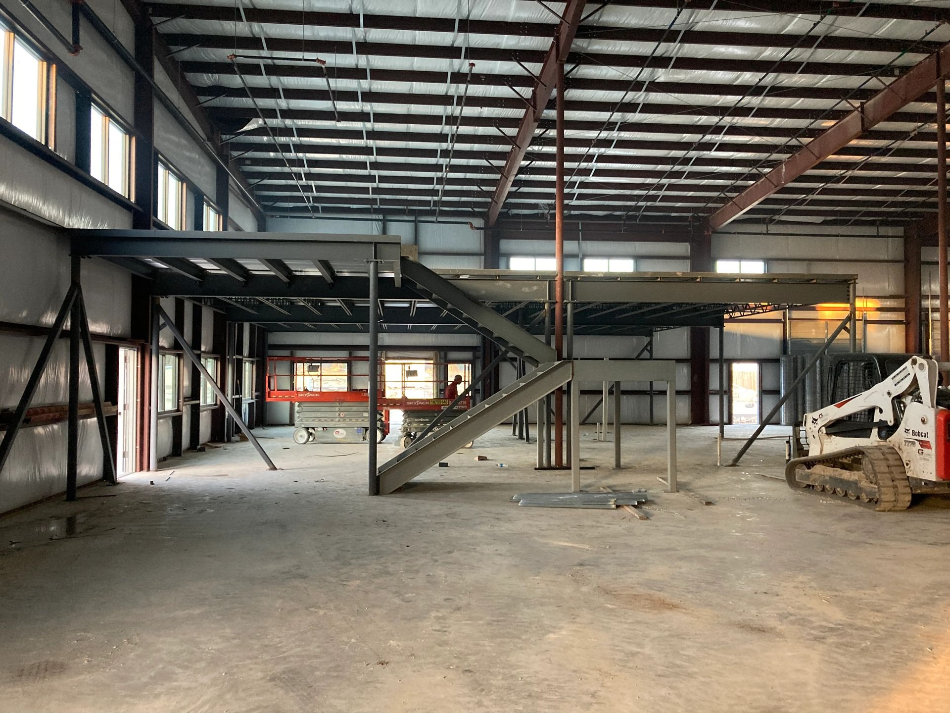 Empty industrial warehouse interior with steel mezzanine, stairs, and a skid-steer loader