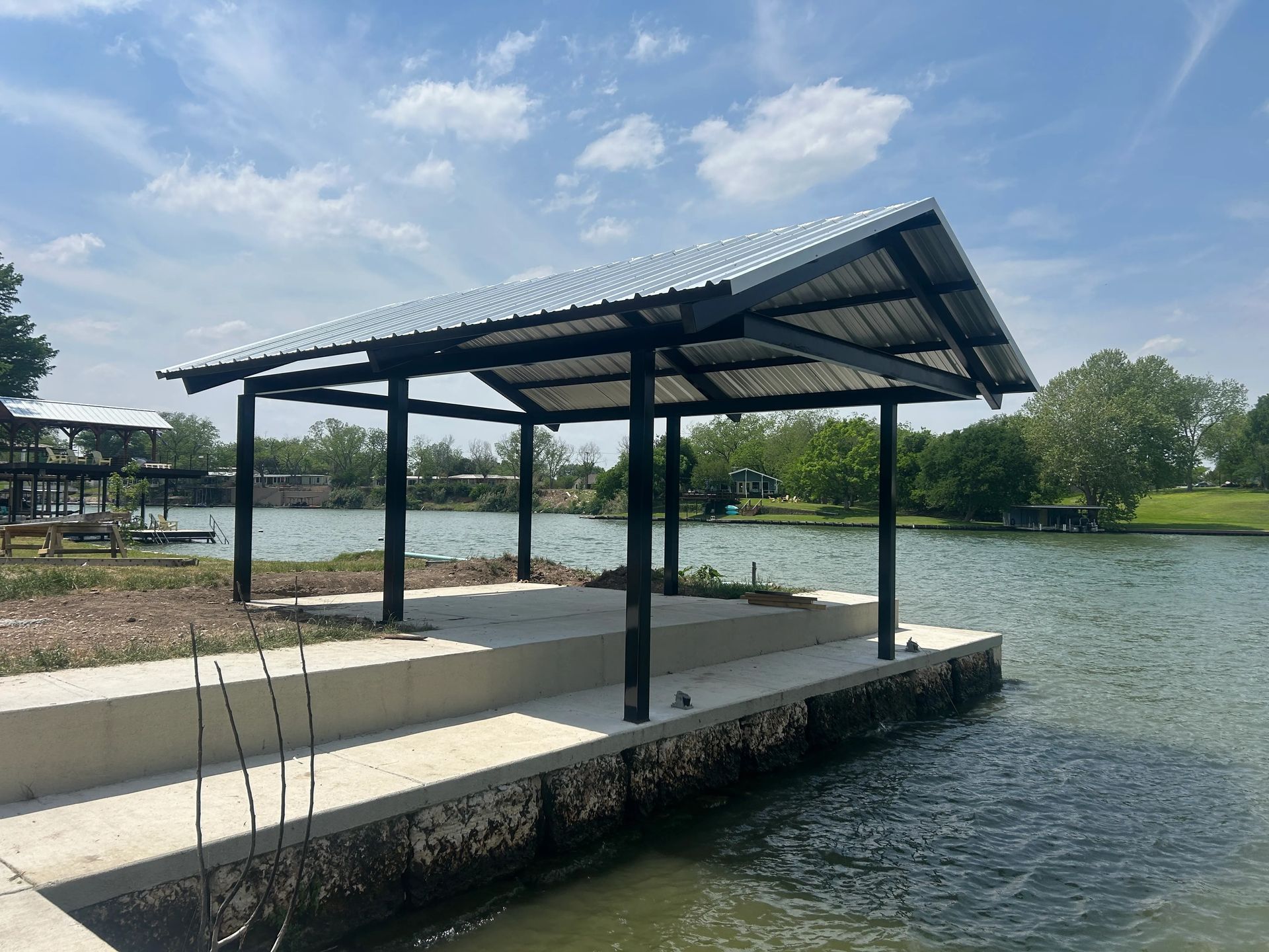 Covered lakeside pavilion with black posts and a metal roof beside calm water under a cloudy sky