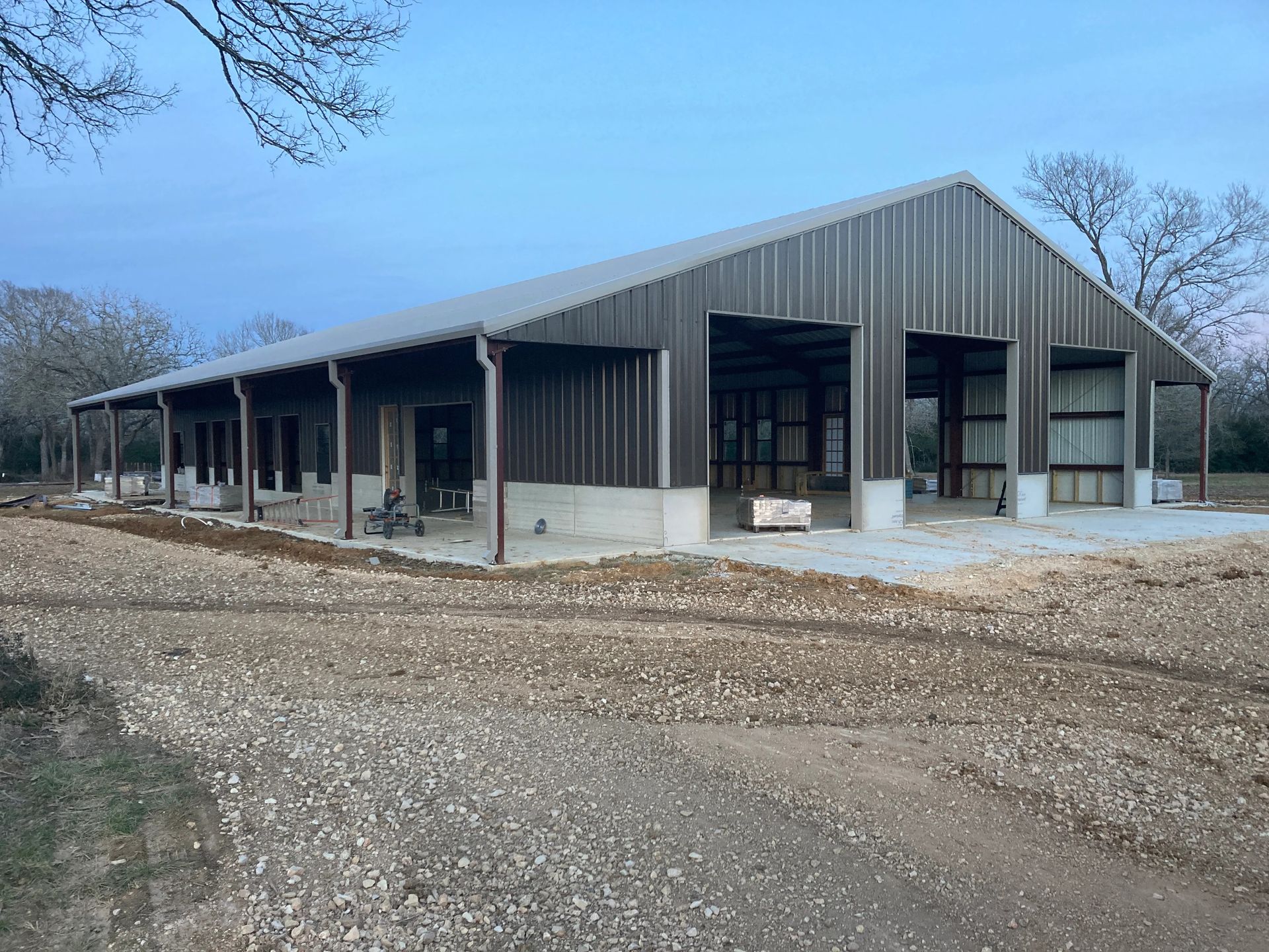 Large barn-style building with a metal roof and open bays beside a gravel road under a blue sky