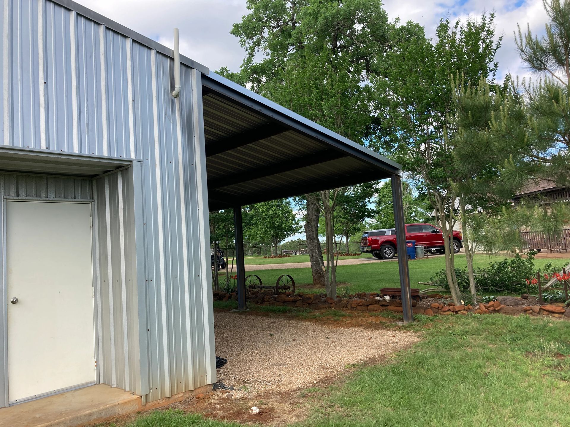 Metal building with a covered carport, gravel path, and red pickup truck in the background