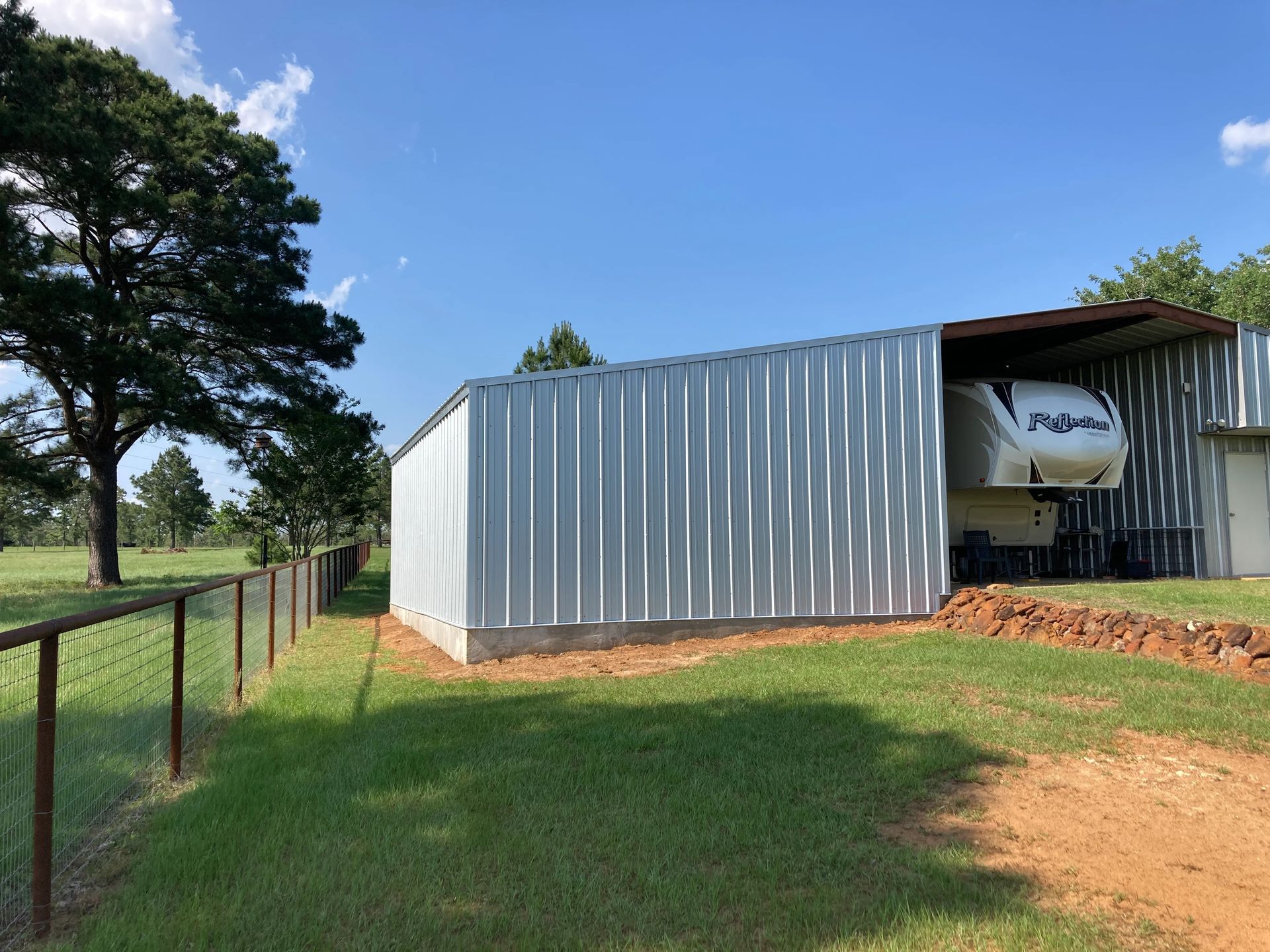 Large metal shed on a grassy rural property, with a white trailer parked inside and trees in the background.